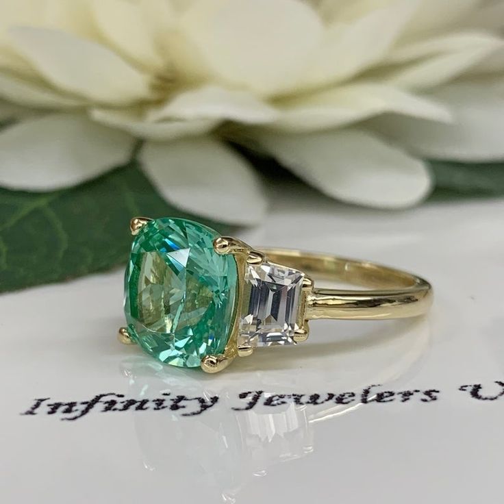 an emerald and diamond ring sits on top of a piece of paper next to a flower