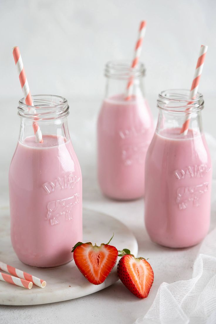 three mason jars filled with pink smoothie and two strawberries on a plate next to each other