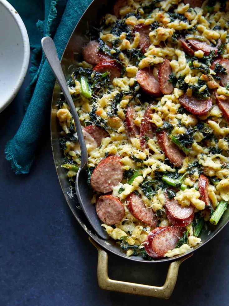 a pan filled with pasta and sausage on top of a blue table cloth next to a white bowl