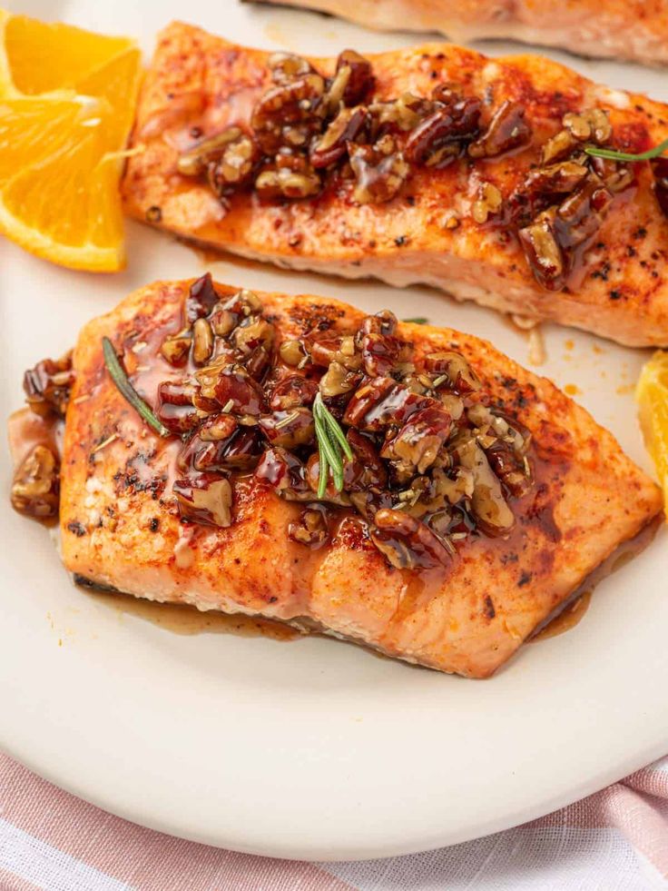 two salmon fillets on a white plate with orange slices and pecans in the background