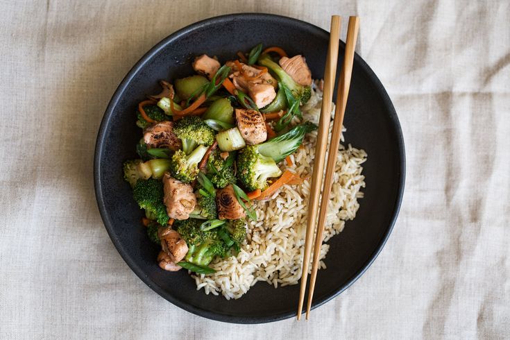 a black plate topped with rice, broccoli and chicken next to chopsticks