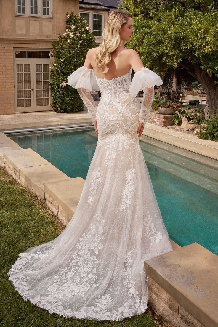 a woman standing in front of a pool wearing a white wedding dress with long sleeves