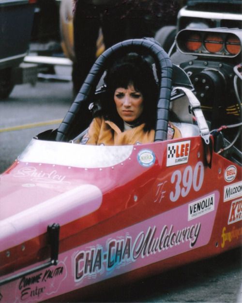 a woman in a pink race car on the street with other cars and people behind her