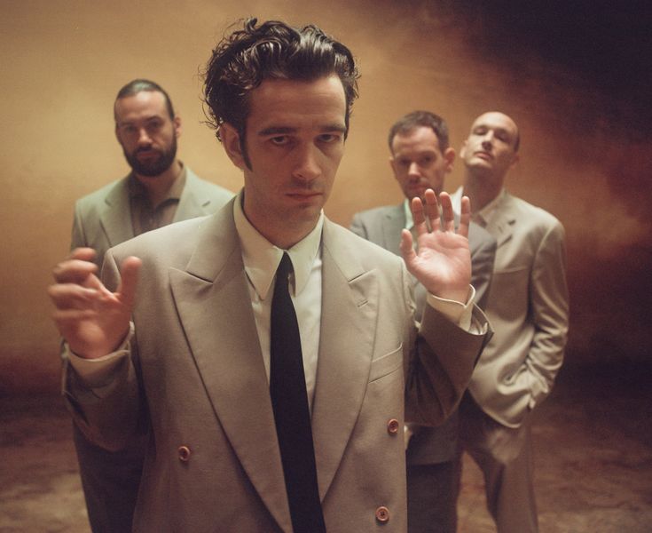 a man in a suit and tie holding his hand up to the camera while three other men stand behind him