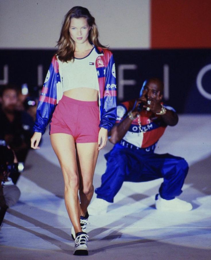 a model walks down the runway with another man behind her wearing blue and red clothing