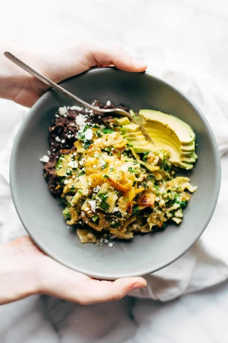 two hands holding a bowl of food with avocado and other foods in it