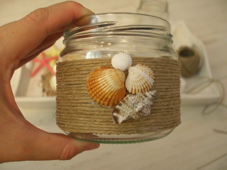 a hand holding a mason jar filled with sea shells and twine wrapped in burlap