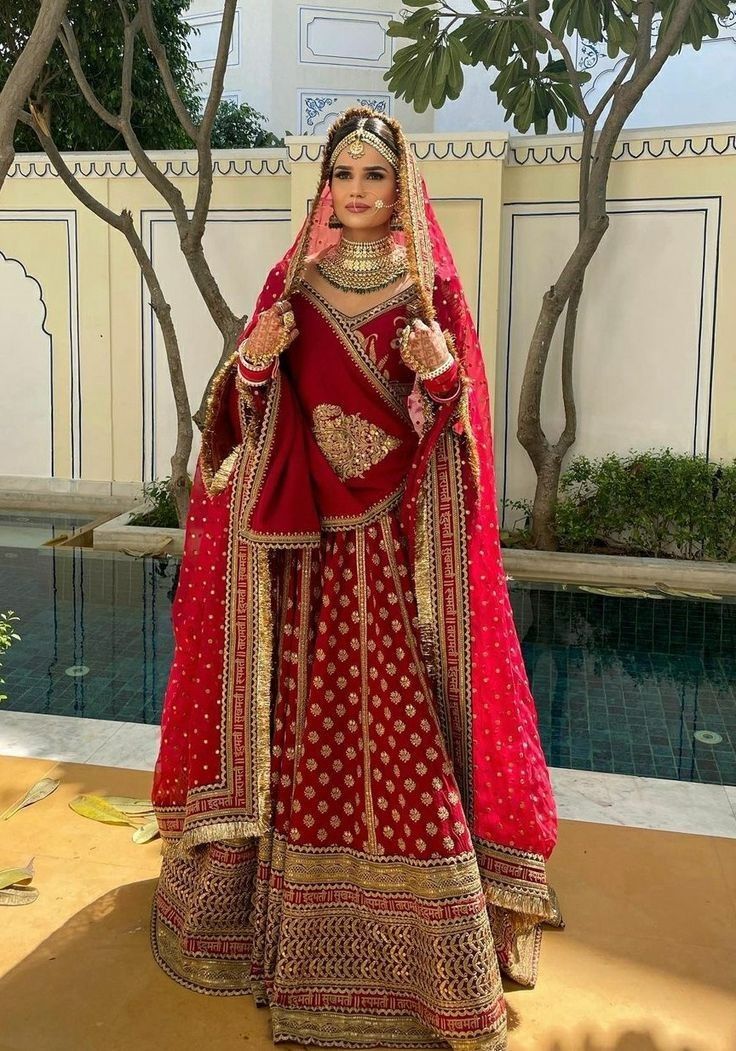 a woman in a red and gold bridal gown standing next to a pool with trees