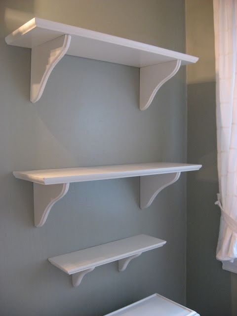 two white shelves on the wall above a toilet in a room with gray walls and curtains