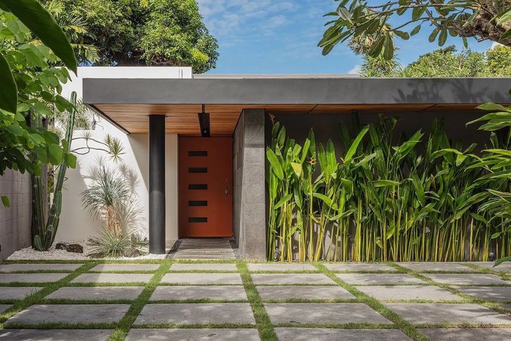 an entrance to a modern home surrounded by lush greenery and bamboo trees, with a red door in the center