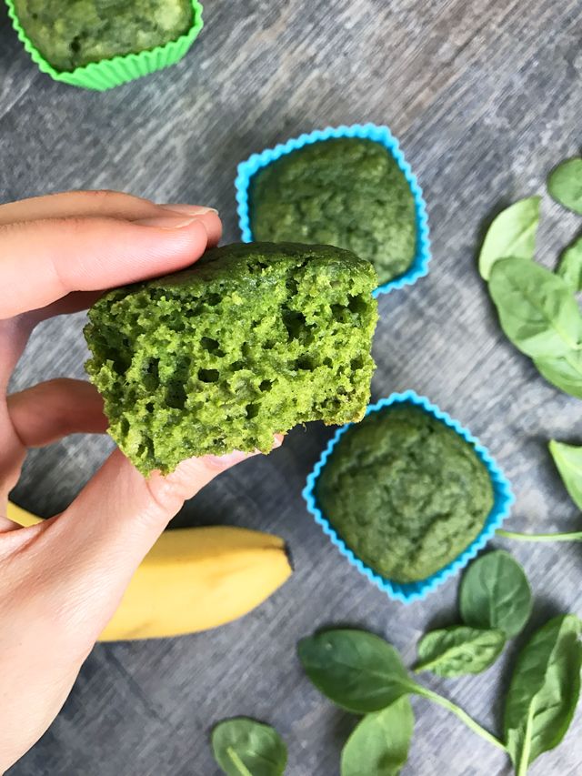 a person holding up a green muffin in front of some bananas and spinach leaves