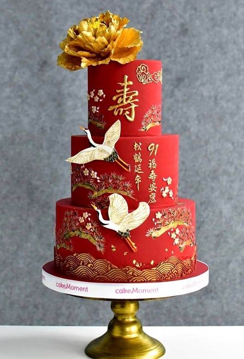 a three tiered red cake with gold decorations on top and flowers in the middle