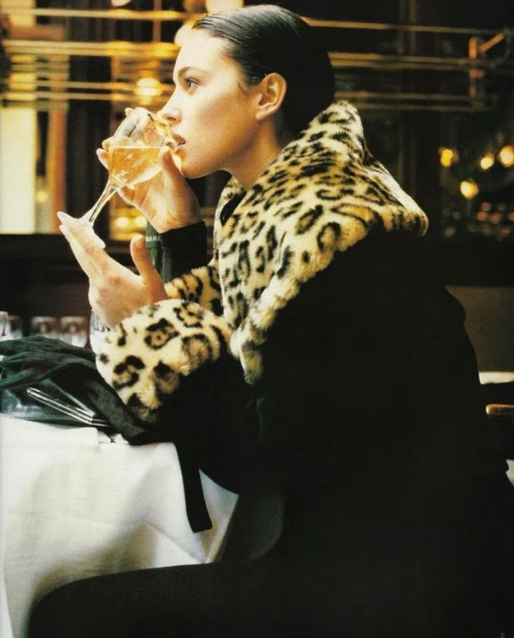 a woman sitting at a table with a wine glass in her hand and wearing a leopard print coat