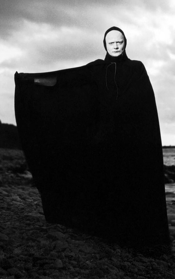 a man dressed in black stands on the beach with his cape draped over his shoulders