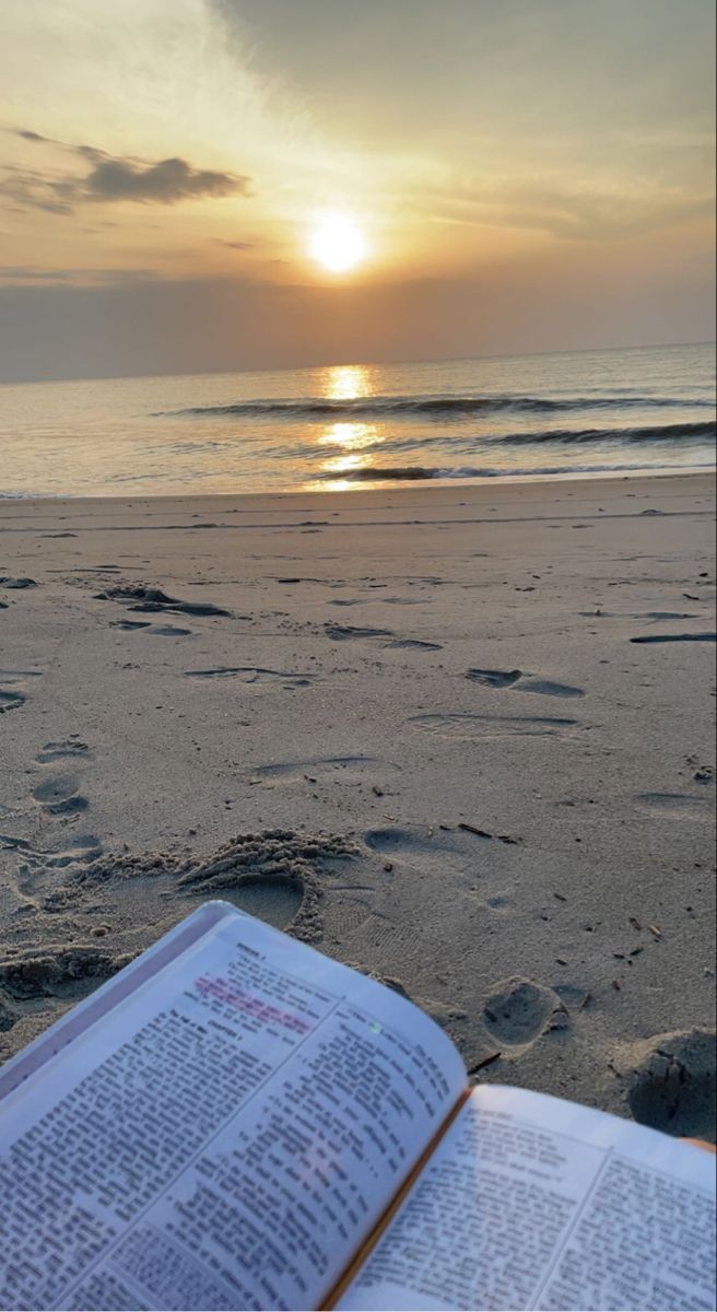 an open book sitting on top of a sandy beach next to the ocean at sunset