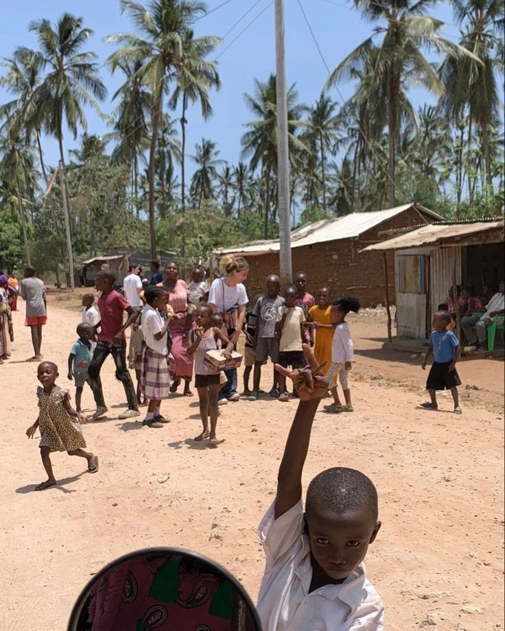 a group of people standing around each other on a dirt road with palm trees in the background