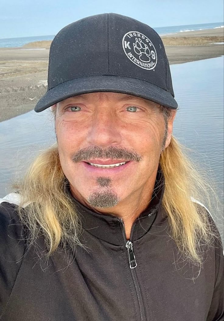 a man with long blonde hair wearing a black hat and smiling at the camera while standing in front of water