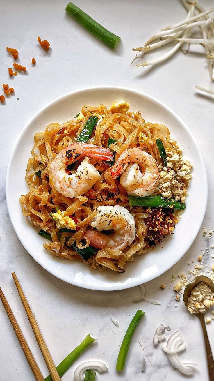 a white plate topped with noodles and shrimp next to chopsticks on a table