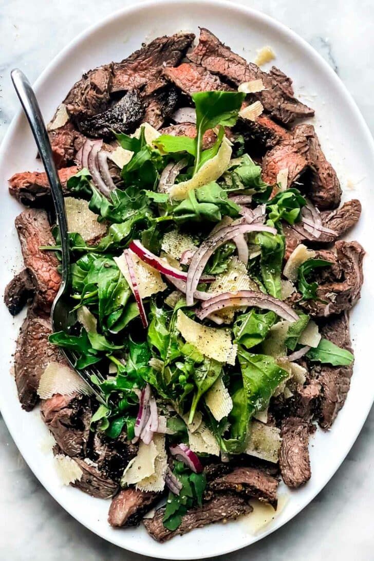 a white plate topped with steak and salad next to a fork on a marble table