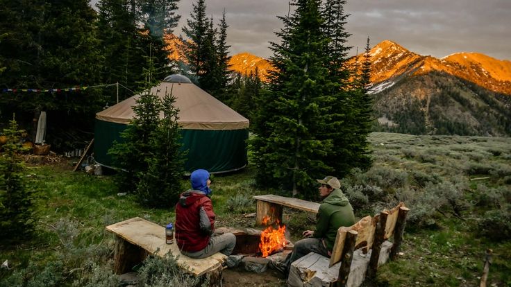 two people sitting around a campfire in the woods at night with mountains in the background