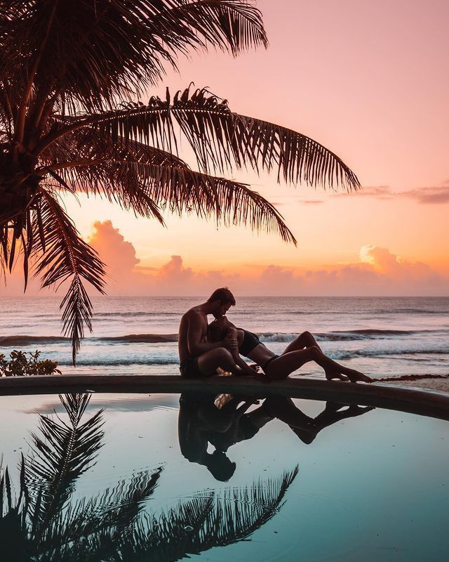 a man sitting on top of a swimming pool next to the ocean at sunset with palm trees