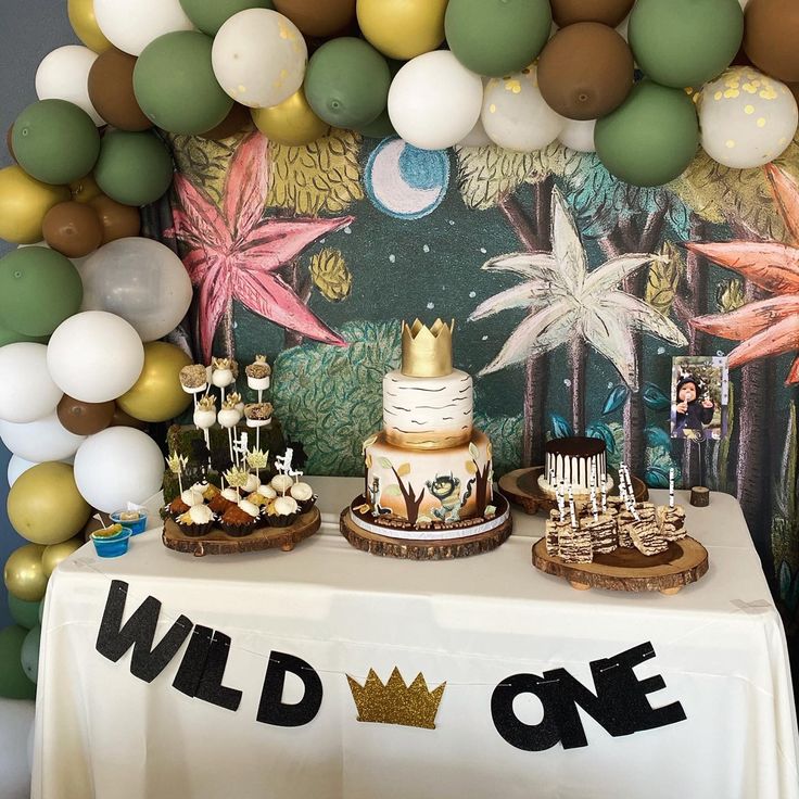 a table topped with cakes and desserts next to a wall covered in balloon garland