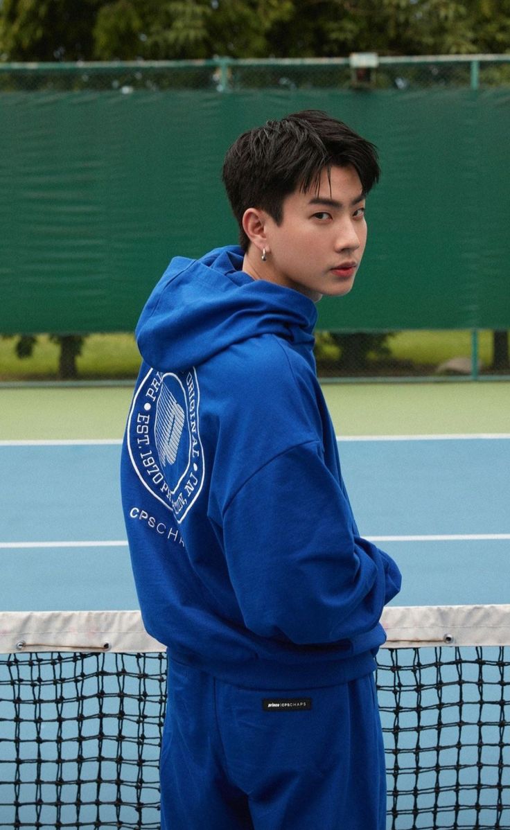 a young man in a blue hoodie stands on a tennis court with his back to the camera