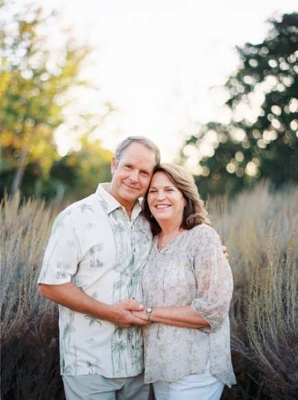 an older man and woman standing in front of tall grass with their arms around each other