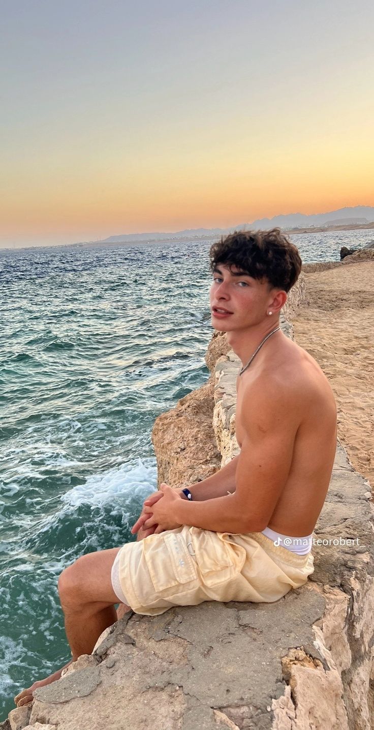 a shirtless man sitting on the edge of a cliff by the ocean at sunset