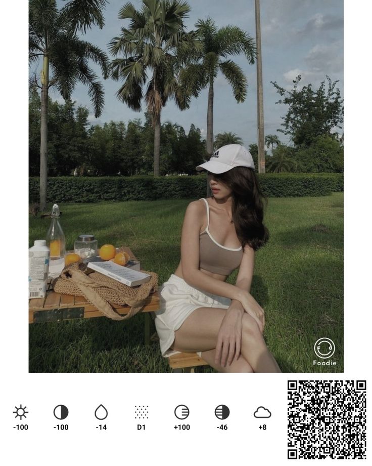 a woman is sitting on the grass near some food and drinks, with palm trees in the background