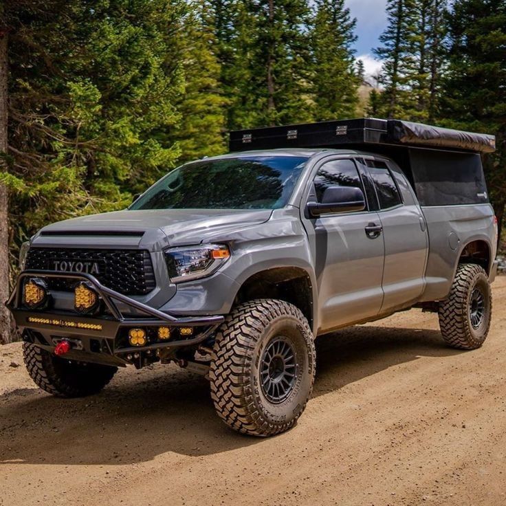 a silver truck is driving on a dirt road in the woods with trees behind it