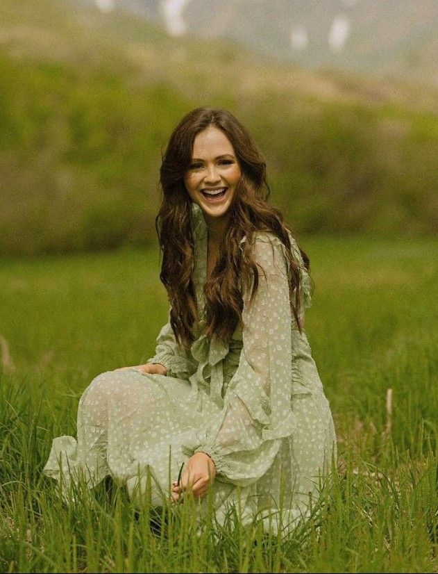 a woman is sitting in the grass and smiling at the camera with mountains in the background