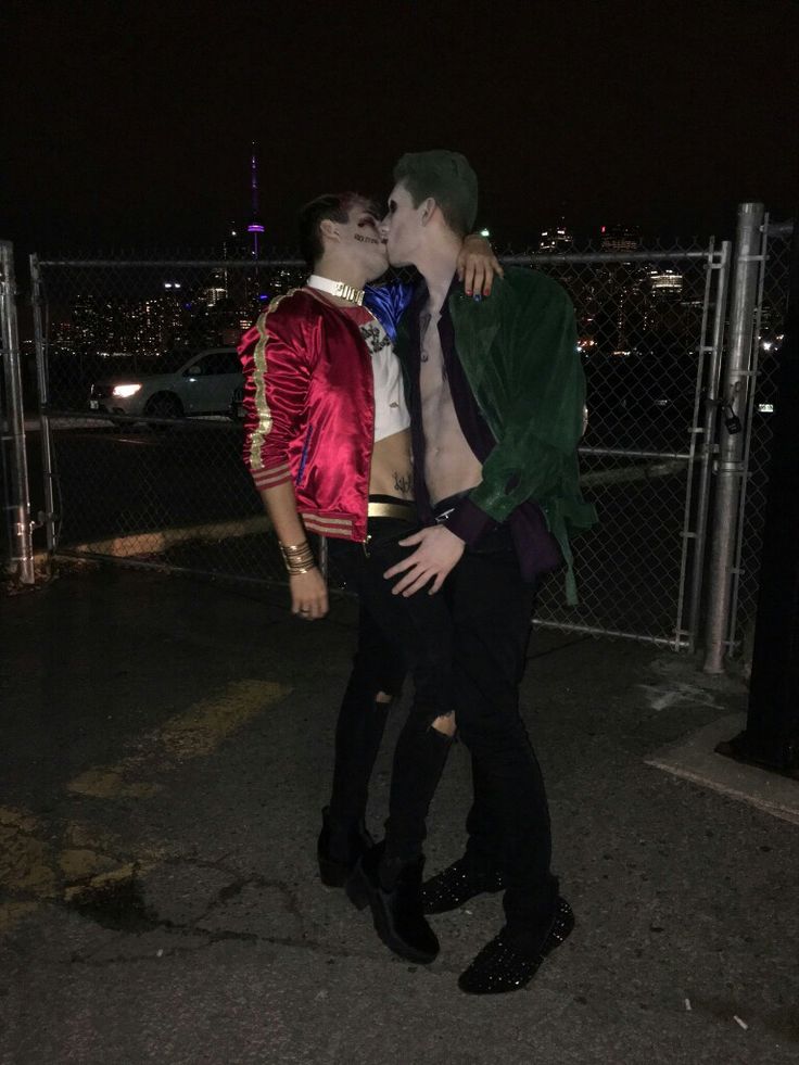 two young men are kissing in front of a fence with the city lights behind them