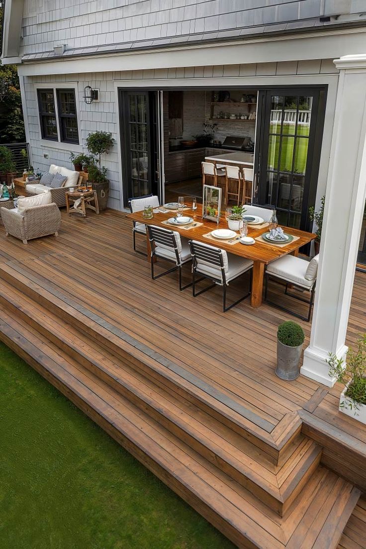 a wooden deck with chairs and table next to a house on the grass covered lawn