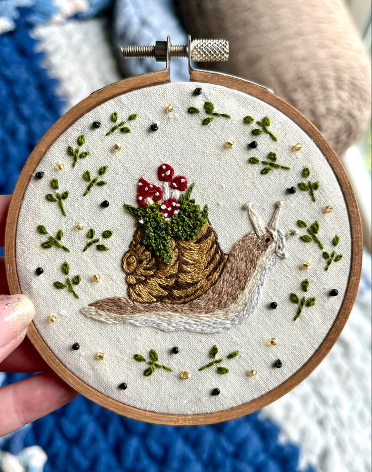 a hand holding a small embroidery hoop with a snail on it's back and some red flowers in the middle