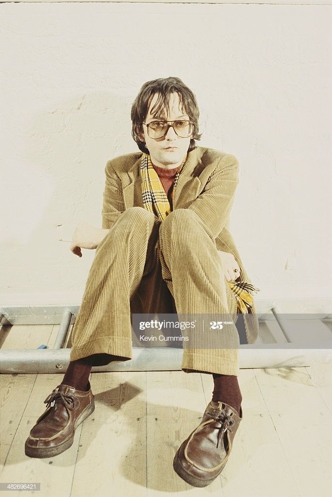 a man sitting on top of a metal bench in front of a white wall wearing glasses