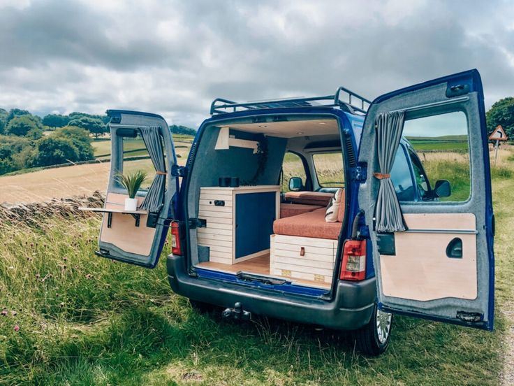 the back end of a van with its doors open on a grassy field next to a stone wall