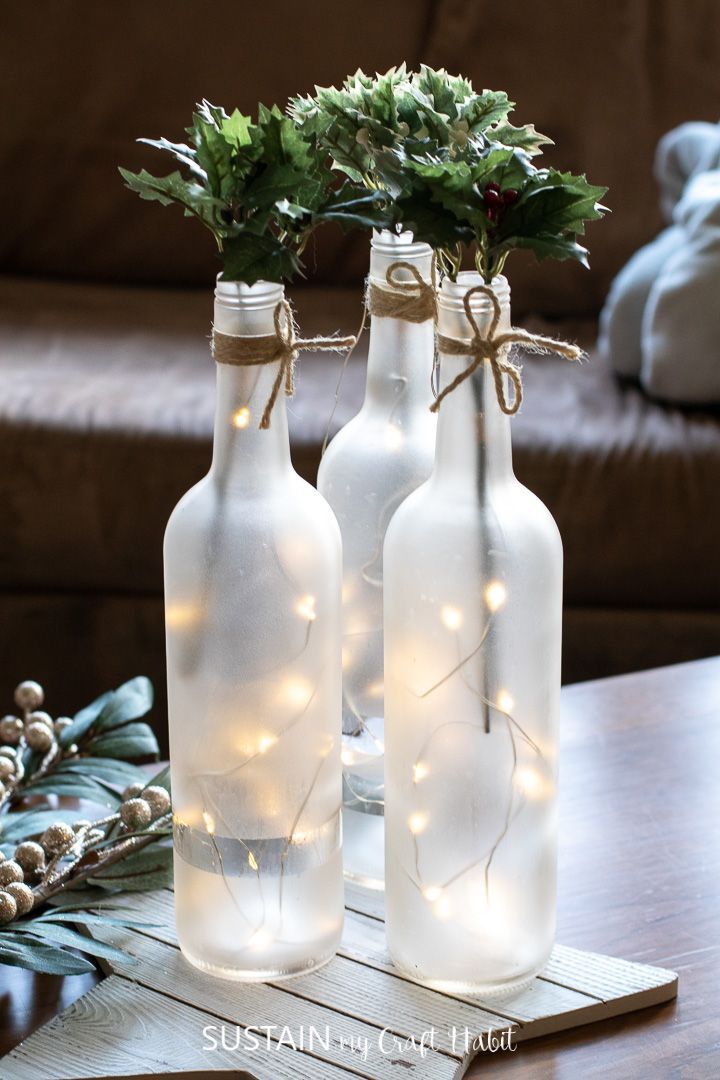 two glass bottles with lights and greenery in them are sitting on a wooden table