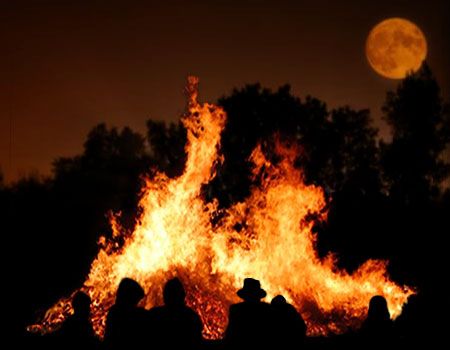 a group of people standing in front of a fire with the moon behind them on a dark night