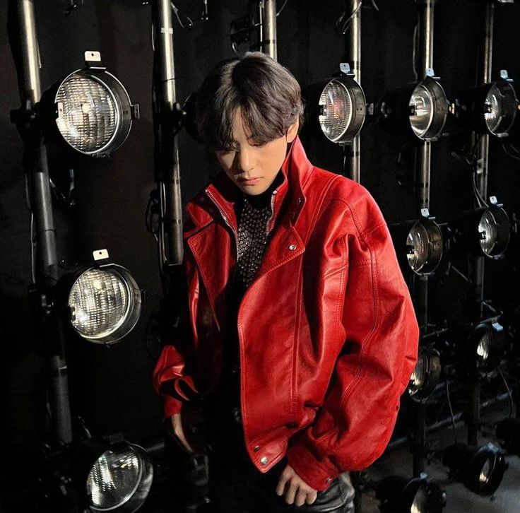 a young man standing in front of a wall of lights wearing a red leather jacket