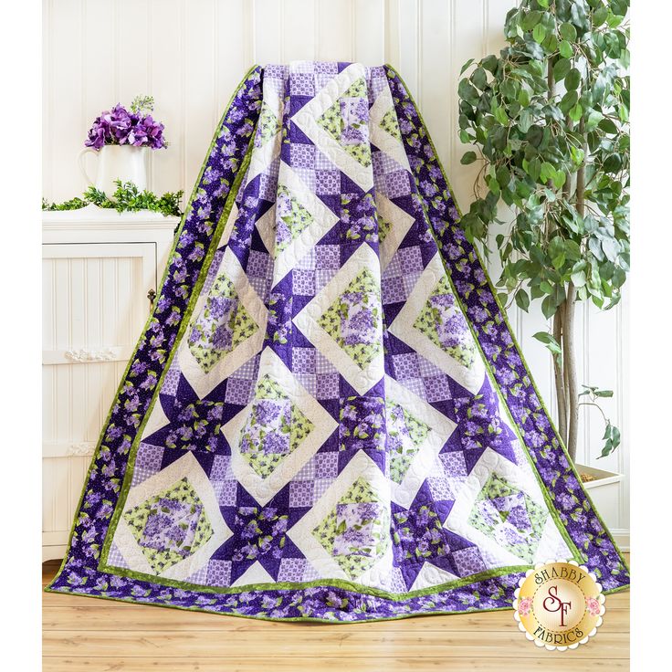 a purple and white quilted blanket sitting on top of a wooden floor next to a potted plant