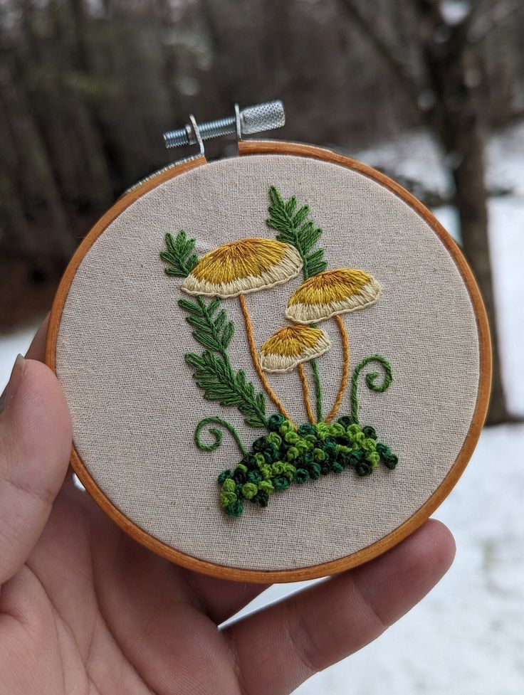 someone is holding up a small embroidered hoop with mushrooms on it and green leaves in the middle