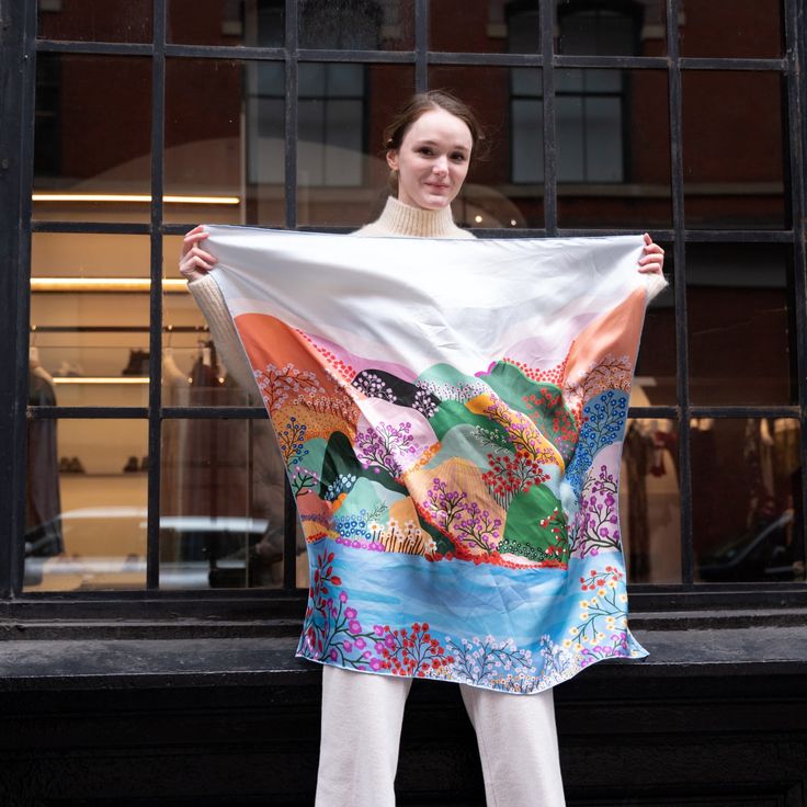 a woman standing in front of a window holding up a large white and multicolored scarf