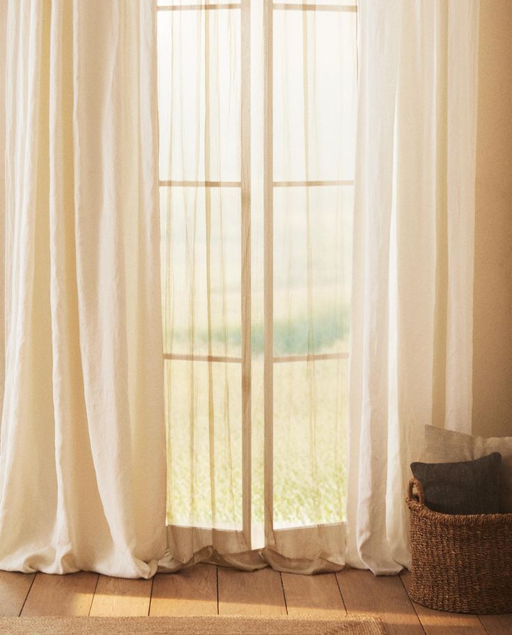 an open window with white curtains and a basket on the floor