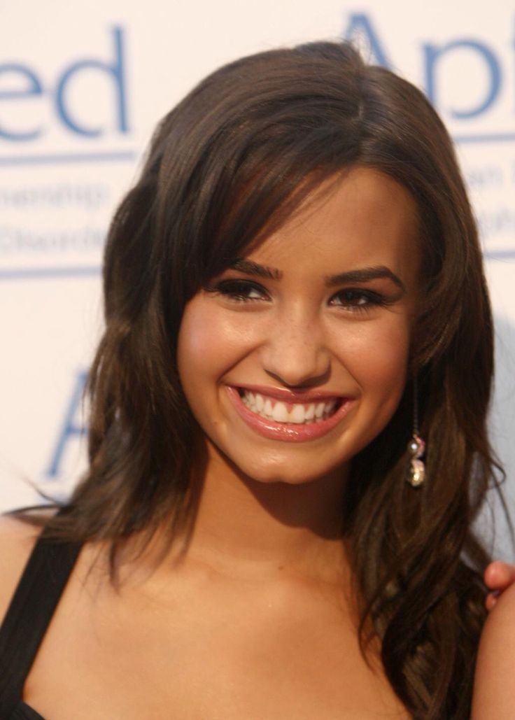 a smiling young woman with long brown hair wearing a black dress and diamond earrings, posing for the camera