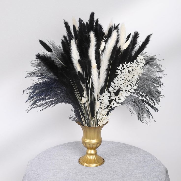 black and white feathers are in a gold vase on a gray tablecloth with a grey background