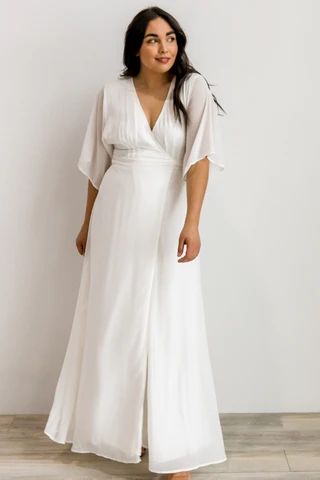 a woman standing in front of a white wall wearing a long white dress and sandals