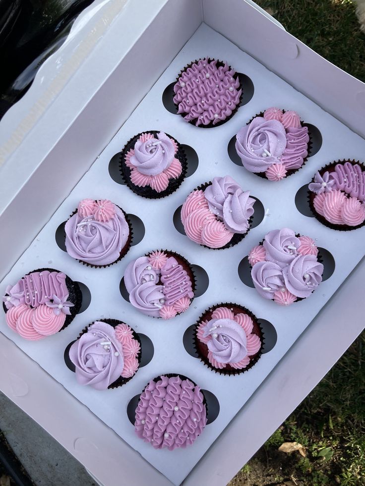 twelve cupcakes with pink and purple frosting in a box on the grass