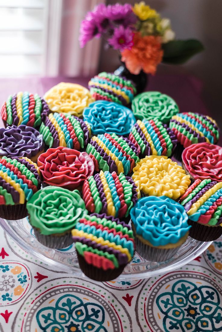 cupcakes decorated with colorful icing are on a glass platter next to flowers