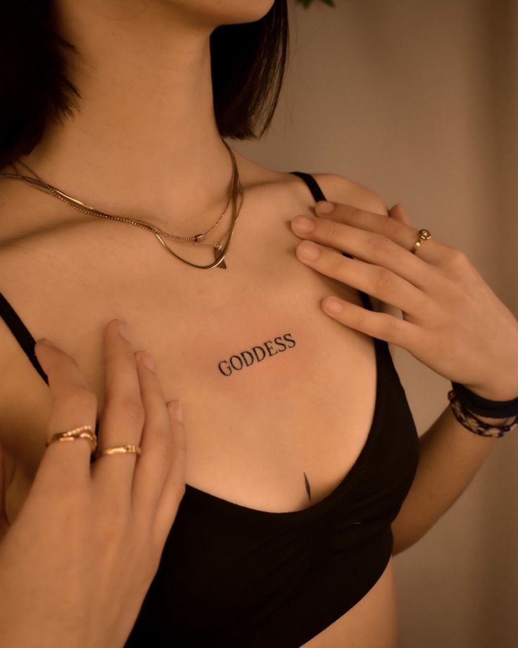 a woman wearing a black bra with the word goddess written on her chest and gold rings